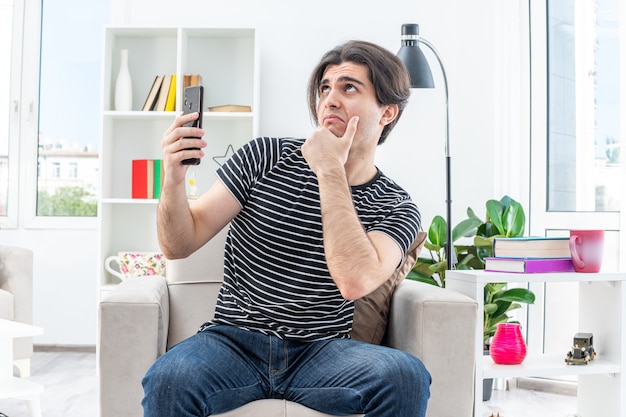 Hombre joven en ropa casual con smartphone mirando hacia arriba con expresión pensativa en la cara pensando sentado en la silla en la sala de luz