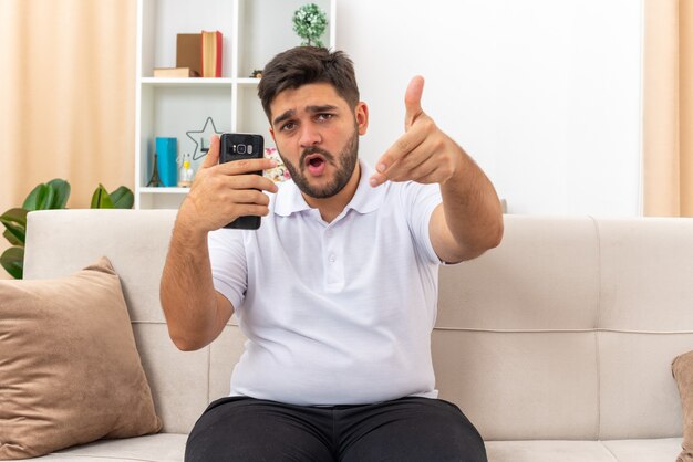 Hombre joven en ropa casual con smartphone mirando feliz y seguro sentado en un sofá en la sala de luz