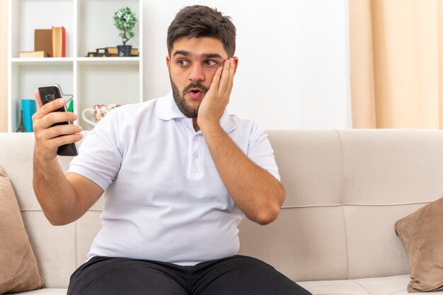 Hombre joven en ropa casual con smartphone mirando la pantalla asombrado y confundido sentado en un sofá en la sala de luz