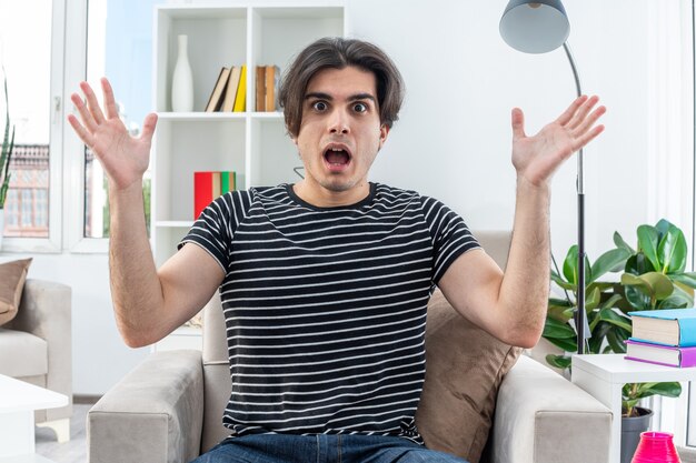 Hombre joven en ropa casual sorprendido y confundido con los brazos levantados sentado en la silla en la sala de estar luminosa