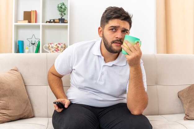 Hombre joven en ropa casual sosteniendo la taza mirando a un lado perplejo pasar el fin de semana en casa sentado en un sofá en la sala de luz