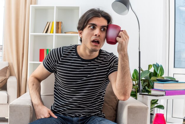 Hombre joven en ropa casual sosteniendo una taza de té mirando preocupado y confundido sentado en la silla en la sala de luz