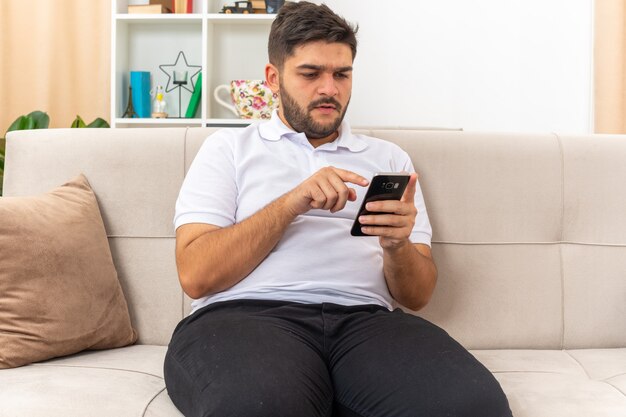 Hombre joven en ropa casual con teléfono inteligente escribiendo varios mensajes mirando la pantalla con cara seria sentado en un sofá en la sala de luz