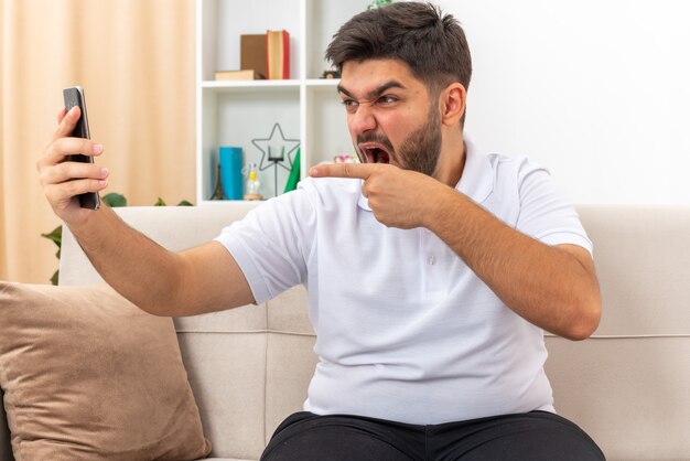 Hombre joven en ropa casual con teléfono inteligente con videollamada enojado y frustrado gritando con expresión agresiva pasando el fin de semana en casa sentado en un sofá en la sala de estar luminosa