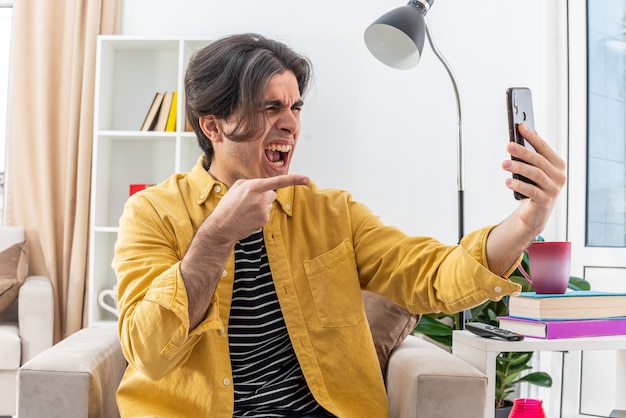 Hombre joven en ropa casual con videollamada con teléfono inteligente enojado y frustrado gritando volviéndose loco sentado en la silla en la sala de estar ligera