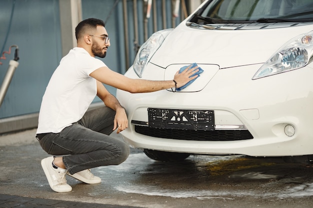 Hombre joven en ropa de moda limpiando su coche con una alfombra después de lavarse en la estación de lavado de coches. Hombre vestido con jeans y camiseta blanca