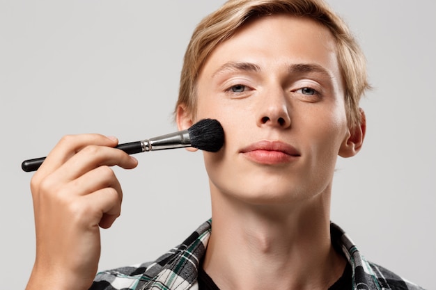 Hombre joven rubio hermoso divertido que lleva la camisa a cuadros casual con el cepillo del maquillaje