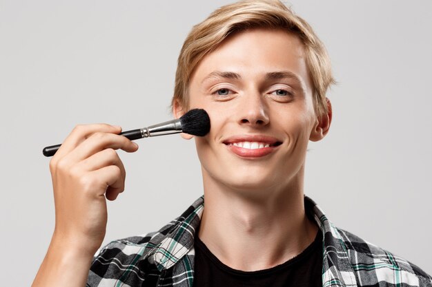 Hombre joven rubio hermoso divertido que lleva la camisa a cuadros casual con el cepillo del maquillaje