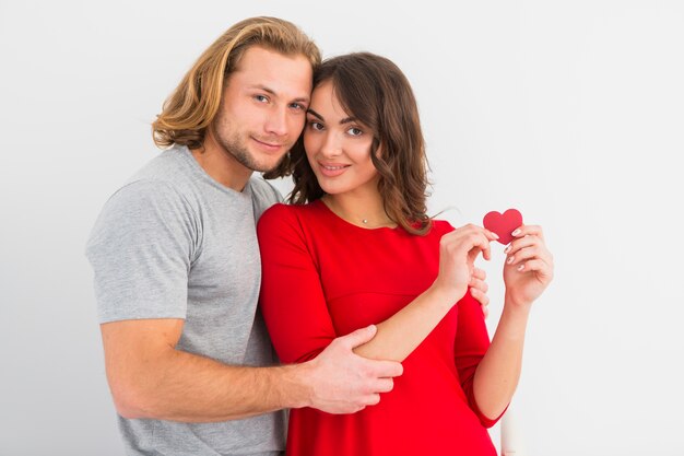 Hombre joven rubio que abraza a su novia sonriente que muestra el papel de la forma del corazón contra el fondo blanco