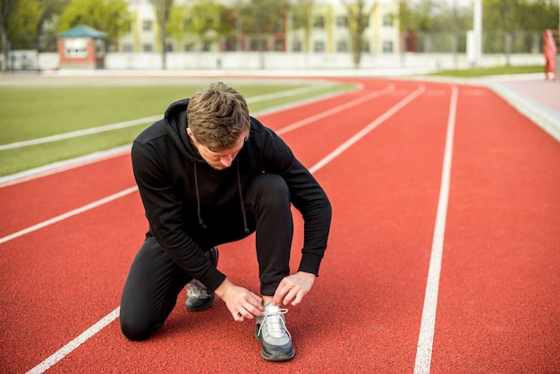 Hombre joven sano sentado en la pista de atletismo atando su cordón
