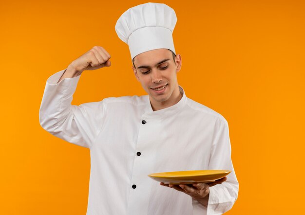 Hombre joven satisfecho fresco vistiendo uniforme de chef mirando la placa mostrando un gesto fuerte