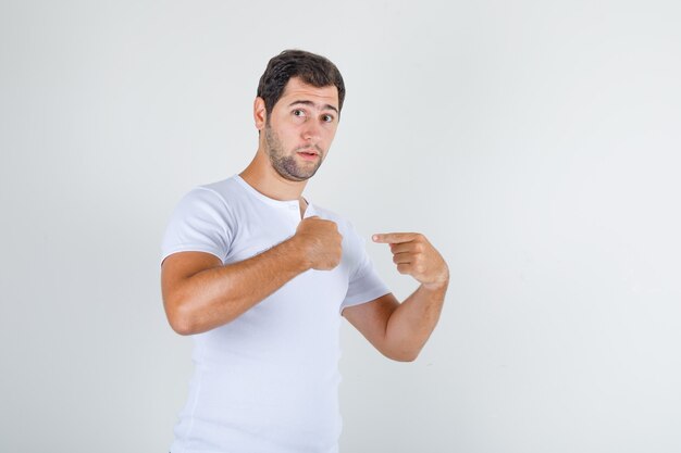 Hombre joven señalando a sí mismo y preguntando "¿yo?" en camiseta blanca, vista frontal.