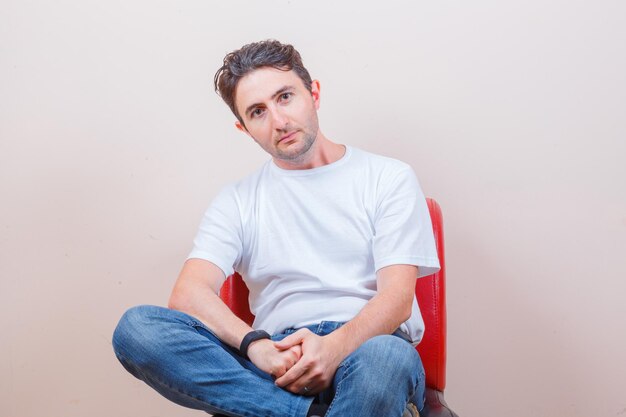 Hombre joven sentado en una silla en camiseta, jeans y mirando confiado