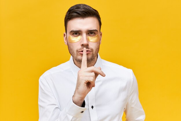 Hombre joven serio con camisa blanca sosteniendo el dedo índice en sus labios, haciendo un gesto de silencio, pidiendo guardar silencio y no decir su secreto, usando parches debajo de los ojos mientras tiene falta de sueño o resaca