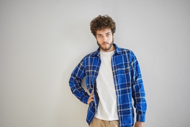 Hombre joven serio elegante aislado con barba espesa y cabello ondulado con mirada segura, sosteniendo la mano en su cintura. Concepto de personas, estilo, moda y ropa masculina.