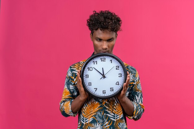 Hombre joven serio y estricto de piel oscura con cabello rizado en hojas camisa estampada sosteniendo reloj de pared que muestra la hora sobre un fondo rosa