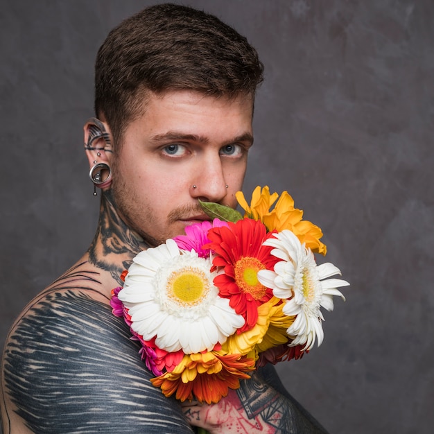 Hombre joven serio con orejas perforadas y nariz sosteniendo una flor de gerbera delante de su boca