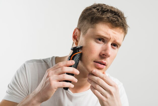 Hombre joven serio que afeita con la máquina contra el fondo blanco