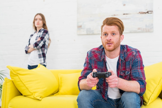 Hombre joven serio que juega al juego con el controlador de video con su novia de pie en el fondo