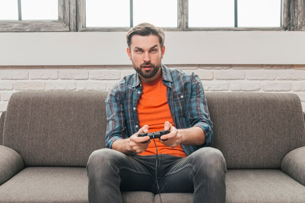 Hombre joven serio que se sienta en el sofá que juega al videojuego