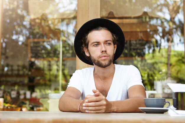 Hombre joven serio con sombrero mirando a distancia con expresión de cara molesta e infeliz mientras está sentado solo en la mesa de café con una taza de té, esperando a su novia