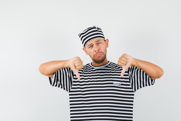Hombre joven con sombrero de camiseta a rayas mostrando doble pulgar hacia abajo y mirando decepcionado