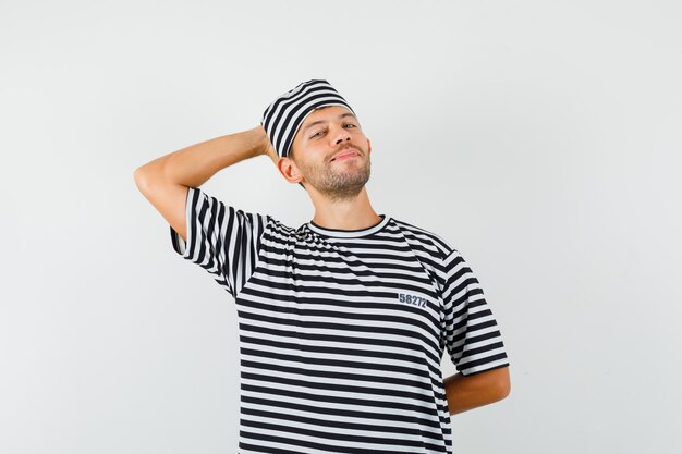 Hombre joven con sombrero de camiseta a rayas sosteniendo la mano detrás de la cabeza y mirando relajado