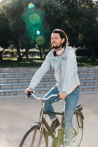 Foto gratuita hombre joven sonriente con los auriculares alrededor de su bicicleta del montar a caballo del cuello en el parque