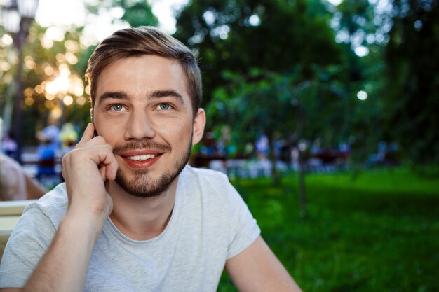 Hombre joven sonriente hermoso que se sienta en la tabla en café al aire libre que habla en el teléfono que mira para arriba.