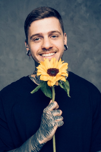 Foto gratuita hombre joven sonriente con las orejas y la nariz perforadas que sostienen el girasol en la mano que mira a la cámara