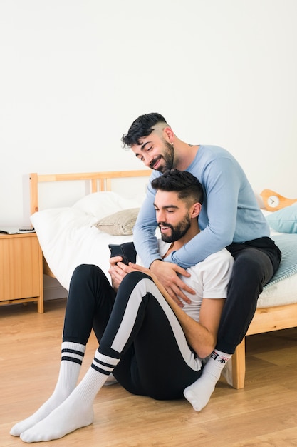 Hombre joven sonriente que abraza a su novio que usa el teléfono móvil