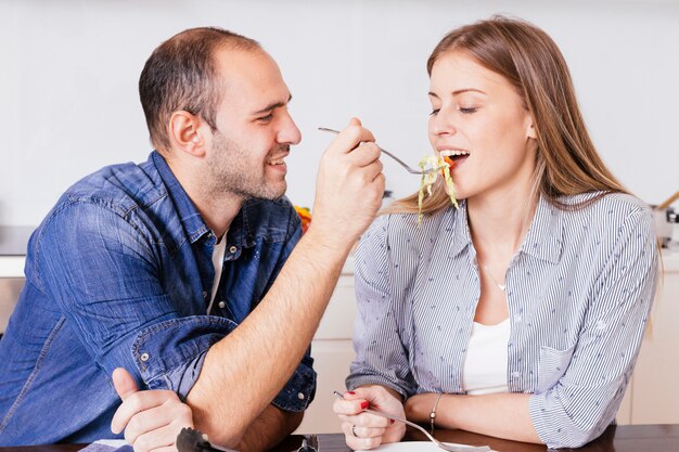 Hombre joven sonriente que alimenta la ensalada a su esposa con la cuchara