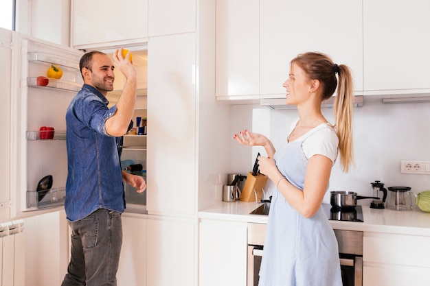 Hombre joven sonriente que se coloca cerca del refrigerador abierto que lanza la verdura en la mano de su esposa