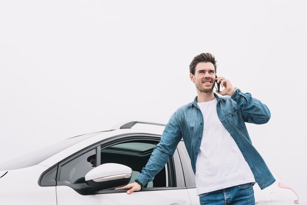 Hombre joven sonriente que se coloca delante del coche que habla en smartphone