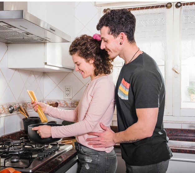 Hombre joven sonriente que está parado detrás de su esposa que prepara espaguetis en casa