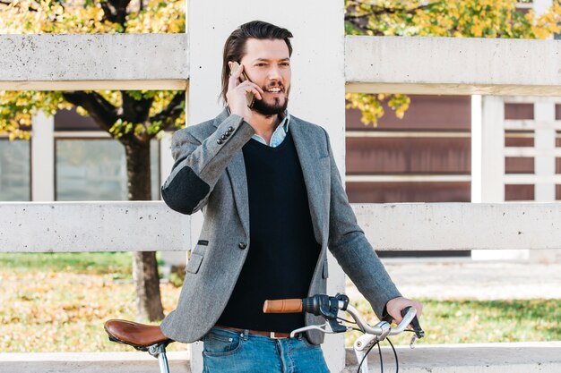 Hombre joven sonriente que habla en el teléfono móvil que se coloca con la bicicleta en el parque