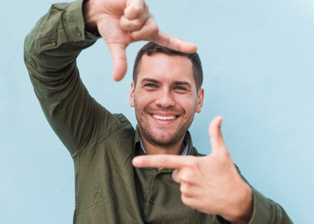 Hombre joven sonriente que hace el marco de la mano sobre fondo azul