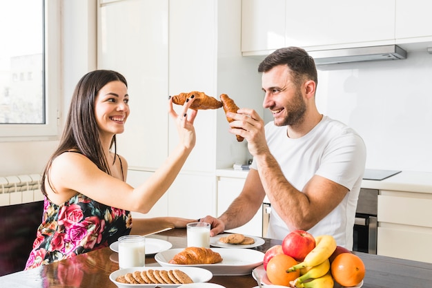 Hombre joven sonriente que juega con el croissant en la cocina