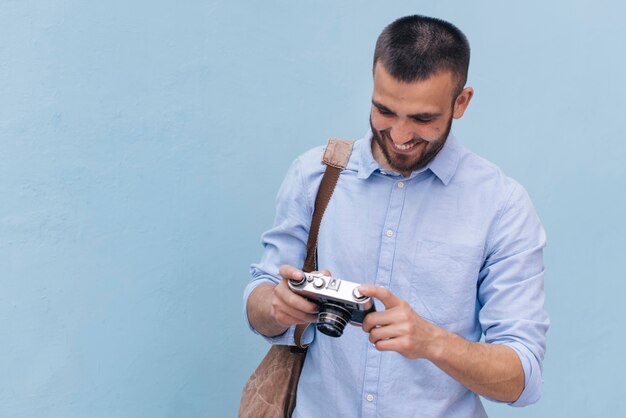 Hombre joven sonriente que lleva la mochila y que mira la cámara que se coloca cerca de la pared azul