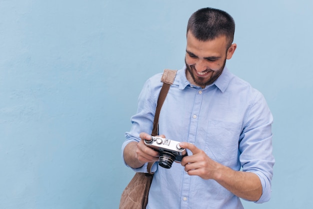 Hombre joven sonriente que lleva la mochila y que mira la cámara que se coloca cerca de la pared azul