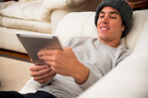 Hombre joven sonriente que miente en el sofá que mira la tableta digital