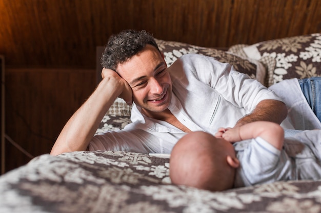 Hombre joven sonriente que miente con su bebé en cama