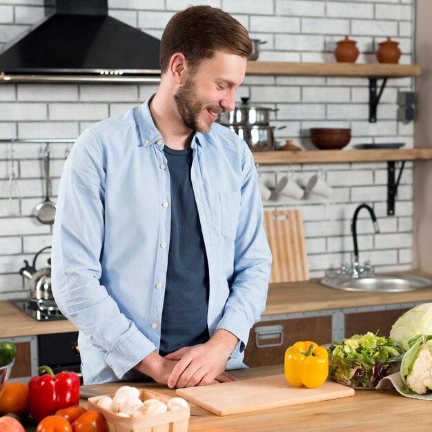 Hombre joven sonriente que mira verduras frescas en contador de la cocina