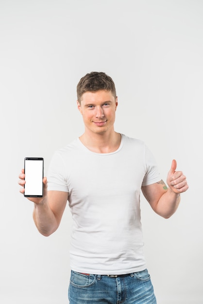 Hombre joven sonriente que muestra el pulgar encima de la muestra que muestra el teléfono elegante contra el fondo blanco