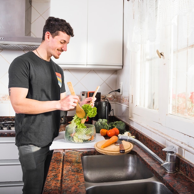 Foto gratuita hombre joven sonriente que prepara la ensalada cerca del fregadero de cocina