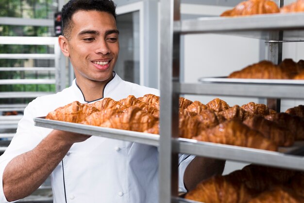 Hombre joven sonriente que quita la bandeja del croissant de la hornada del estante