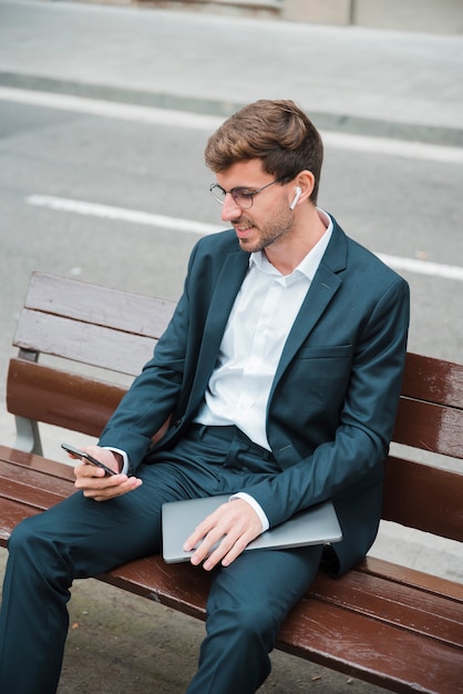 Hombre joven sonriente que se sienta en el banco sobre la calle usando el teléfono móvil con el auricular inalámbrico