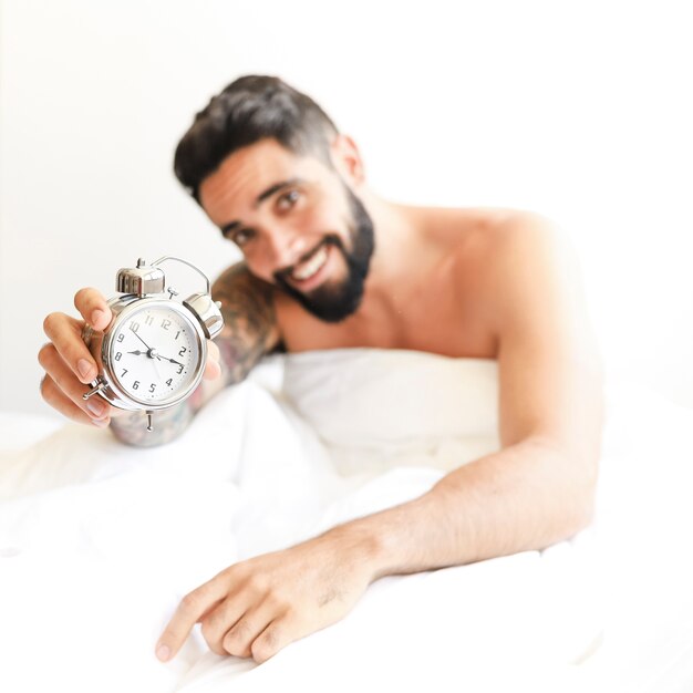 Hombre joven sonriente que se sienta en la cama que muestra el despertador