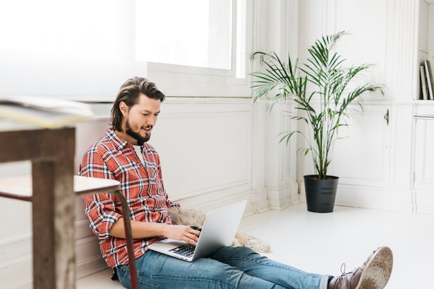 Foto gratuita hombre joven sonriente que se sienta en casa usando el ordenador portátil