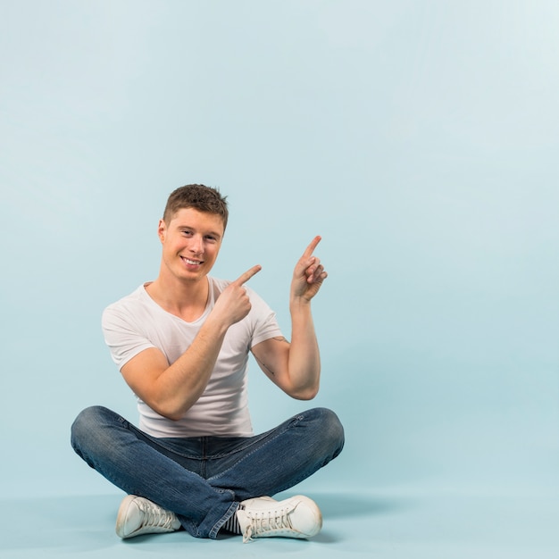 Hombre joven sonriente que se sienta en el piso que señala sus fingeres para arriba contra el contexto azul